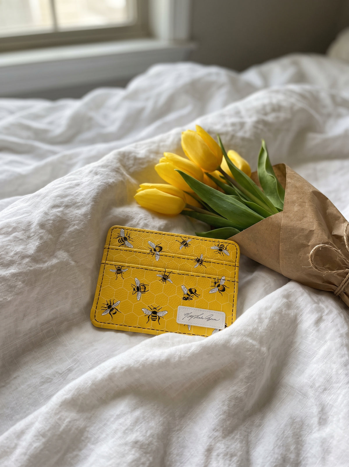 Monarque Mary Lake-Thompson Bees slim wallet card holder on white bedding with yellow tulips